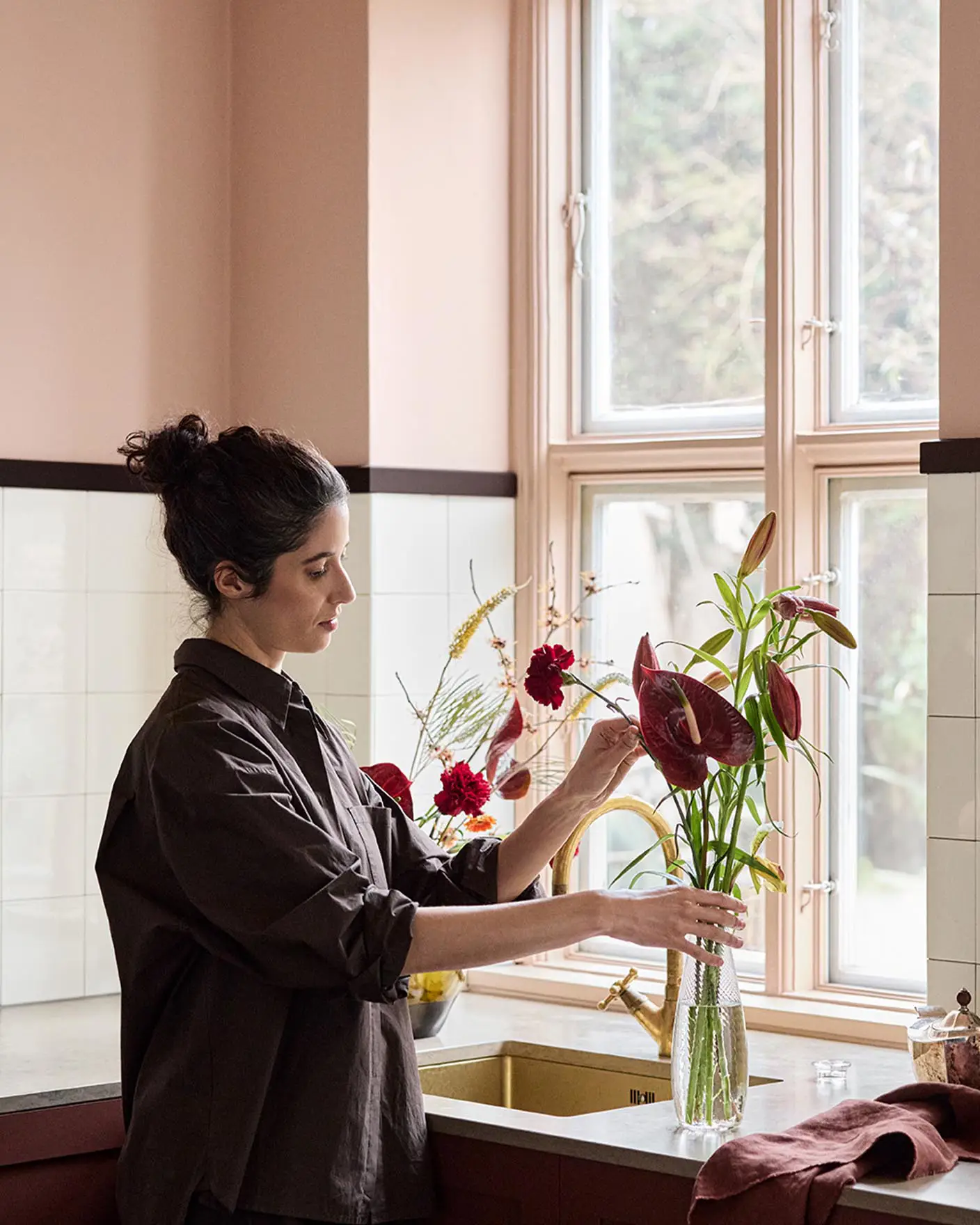 Woman arranging flowers on a kitchen counter with soft pink walls painted with the Jotun colour 20362 Pink Ambience and white tile backsplash.