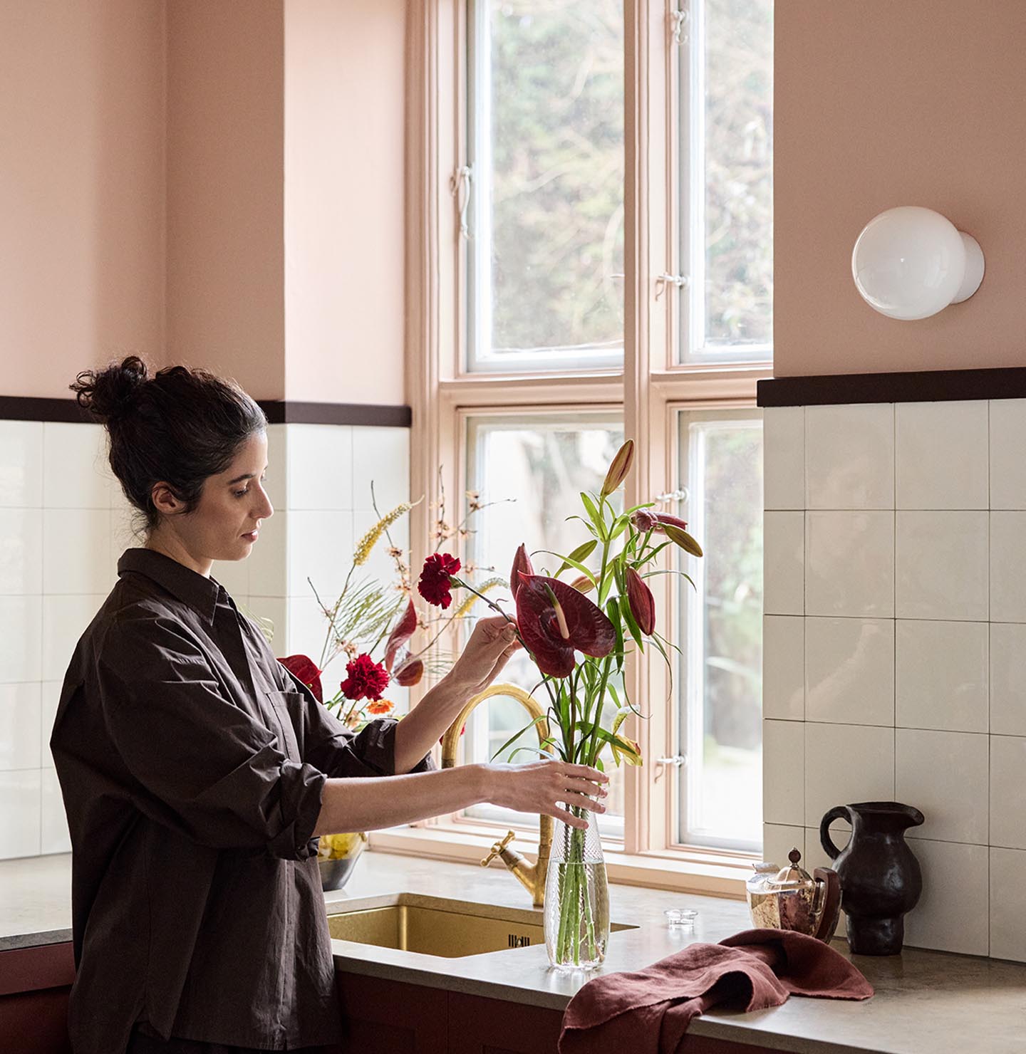 Woman arranging flowers on a kitchen counter with soft pink walls painted with the Jotun colour 20362 Pink Ambience and white tile backsplash.