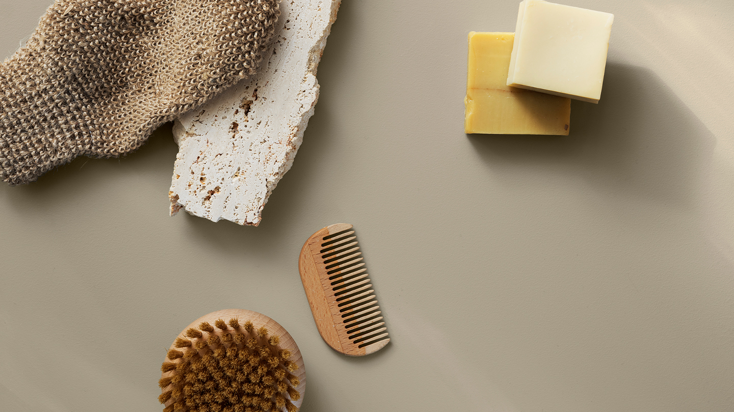 Naturlige badeprodukter på greige overflate: strikket vaskevott, to såpestykker, trekam, skrubbebørste og en rustikk flisstein