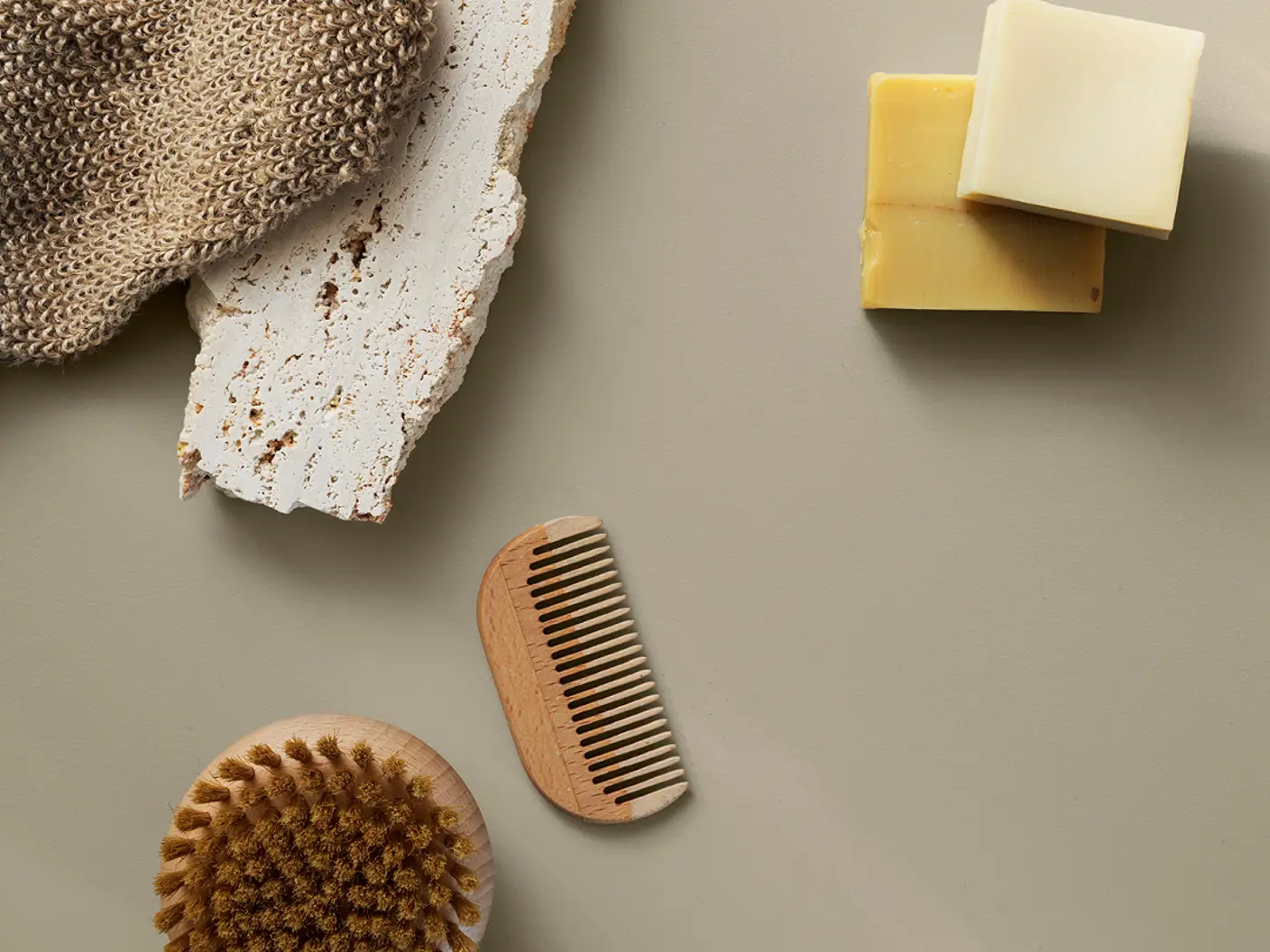 Naturlige badeprodukter på greige overflate: strikket vaskevott, to såpestykker, trekam, skrubbebørste og en rustikk flisstein