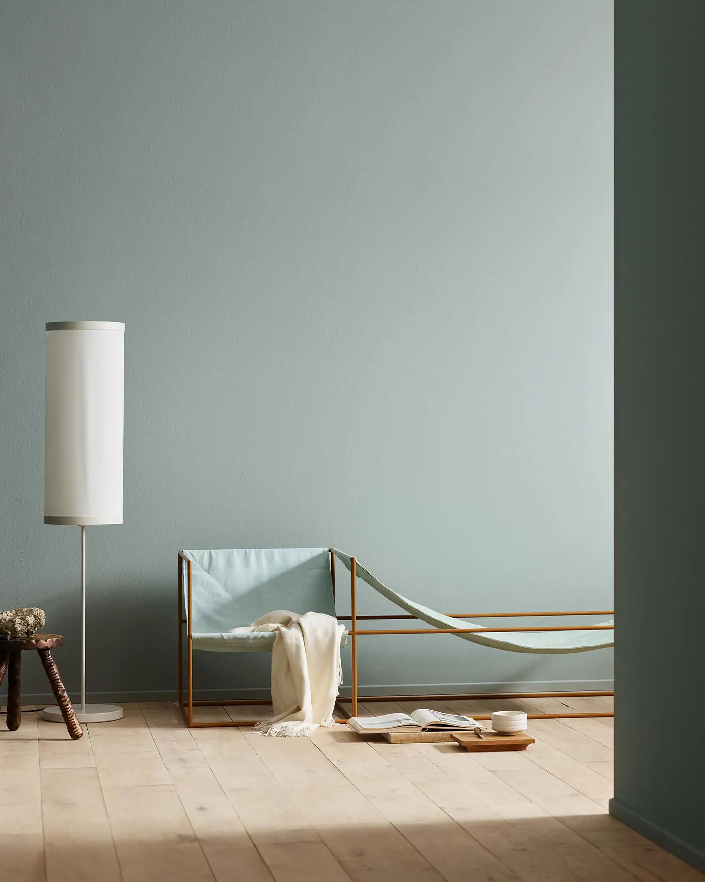 Minimalist bedroom with a light blue chair, white blanket, and wooden stool against a muted blue wall painted with the colour 6378 Iconic from Jotun.