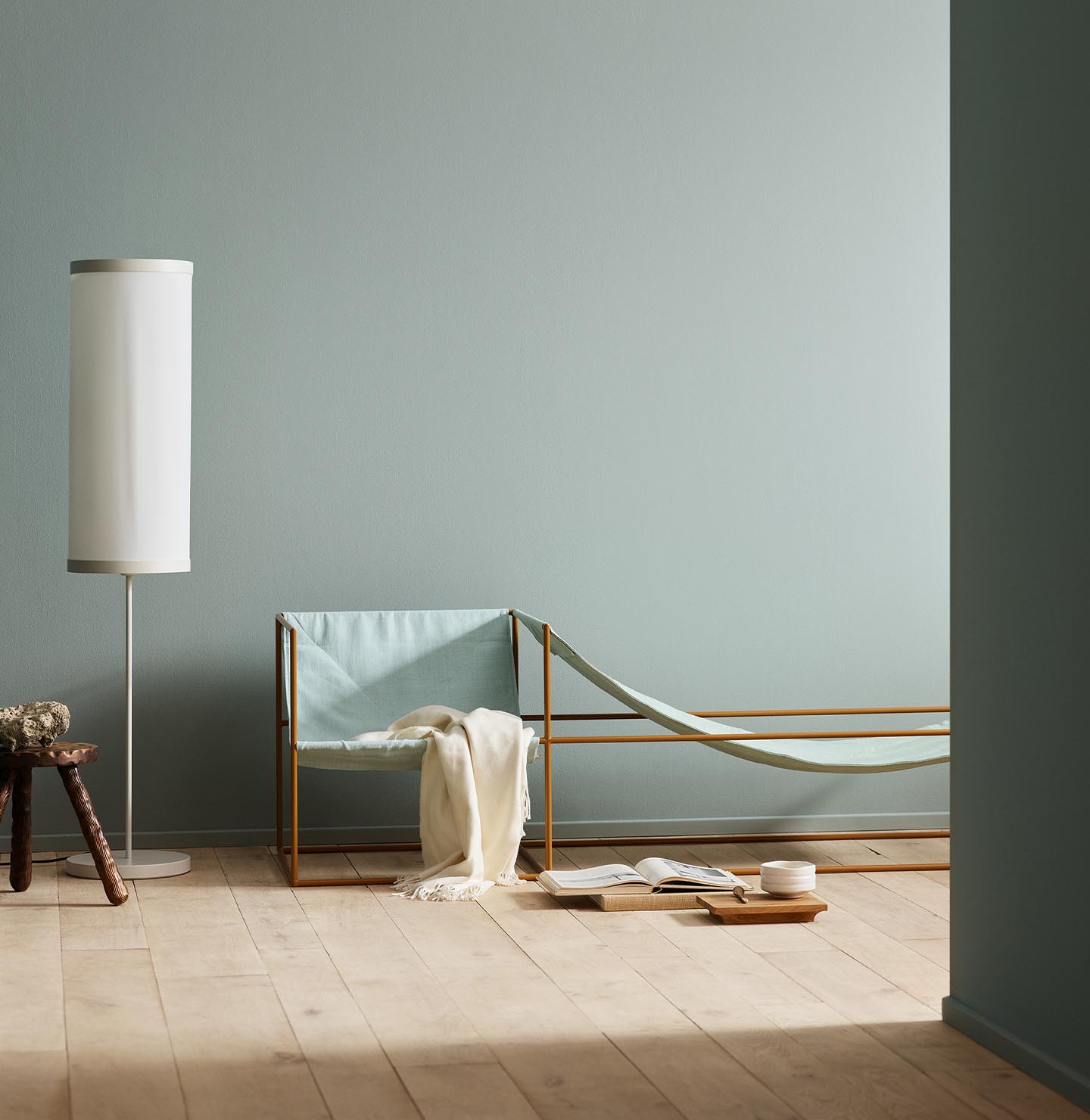 Minimalist bedroom with a light blue chair, white blanket, and wooden stool against a muted blue wall painted with the colour 6378 Iconic from Jotun.