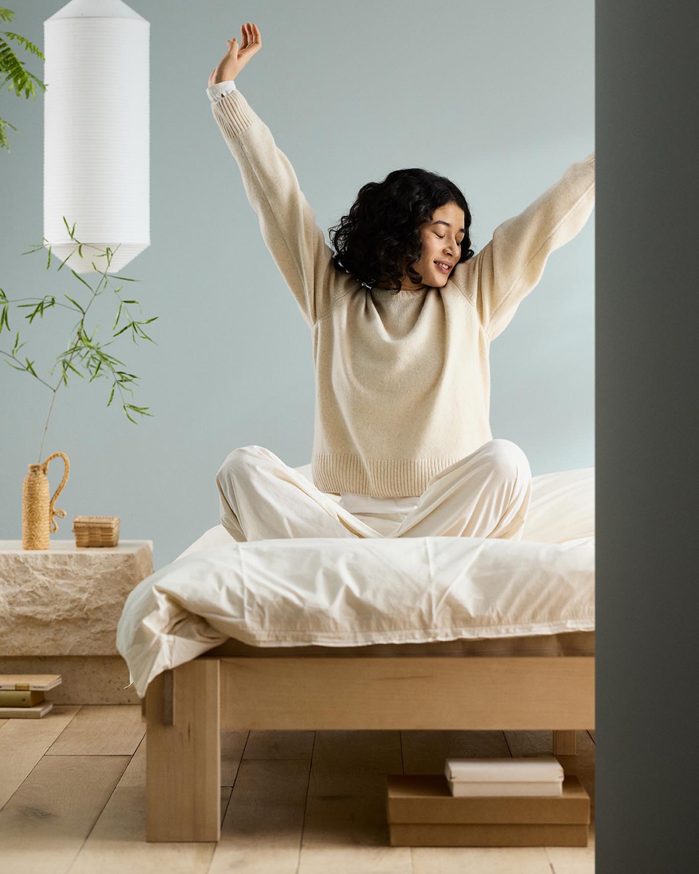 Woman sitting on a bed with arms raised, next to a bedside table against a muted blue wall painted in the Jotun colour 6378 Iconic.