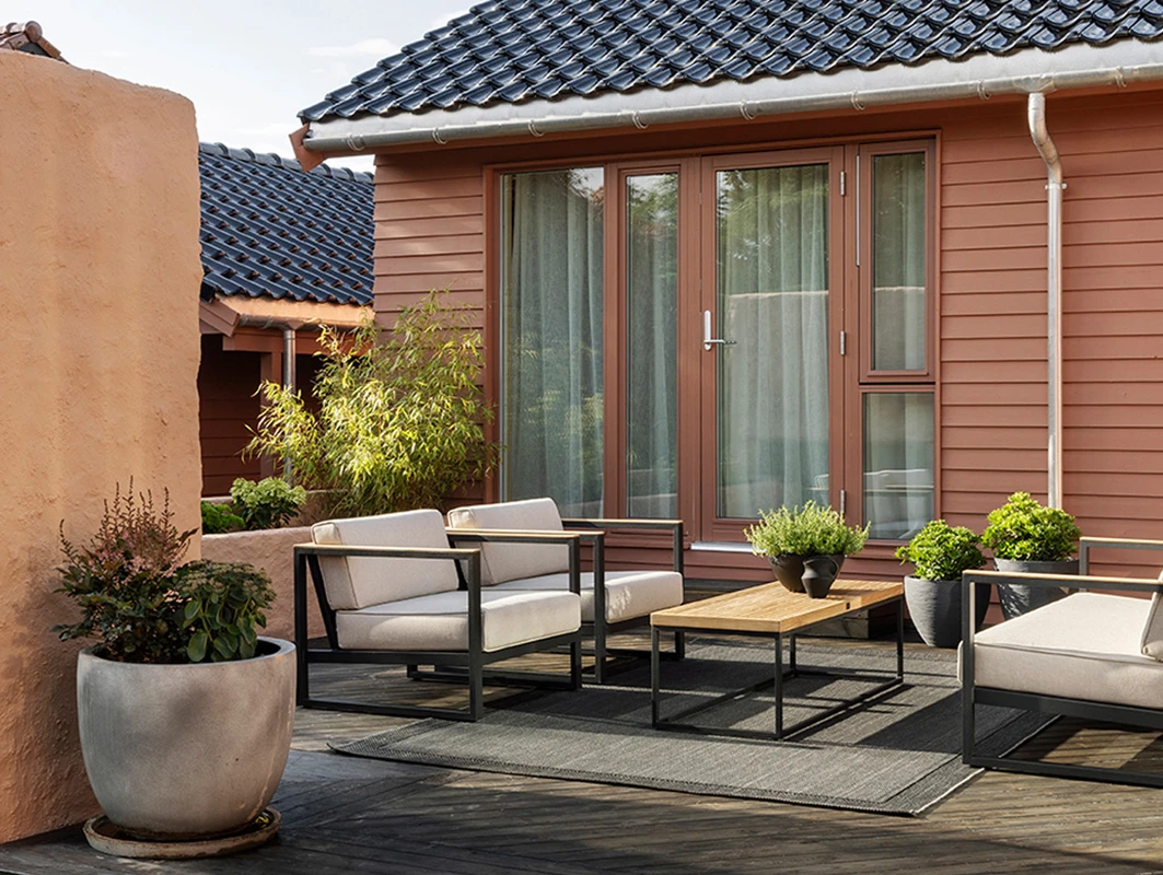 Moderne utemøbler på en terrasse med trepanel, omringet av planter i potter, husveggen er malt lys rød i Jotun 20118 Amber Red