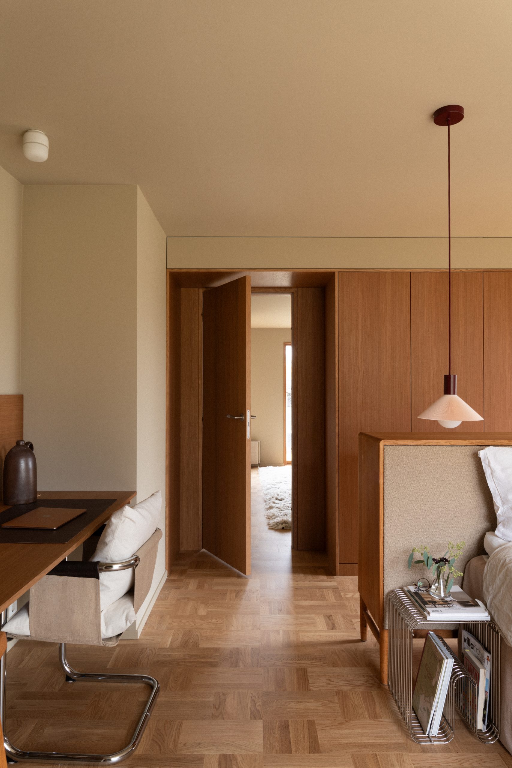 Moderne soverom med tremøbler, åpen dør til annen rom, pendellampe, skrivebord med stol, og sengebenk med dekorative bøker