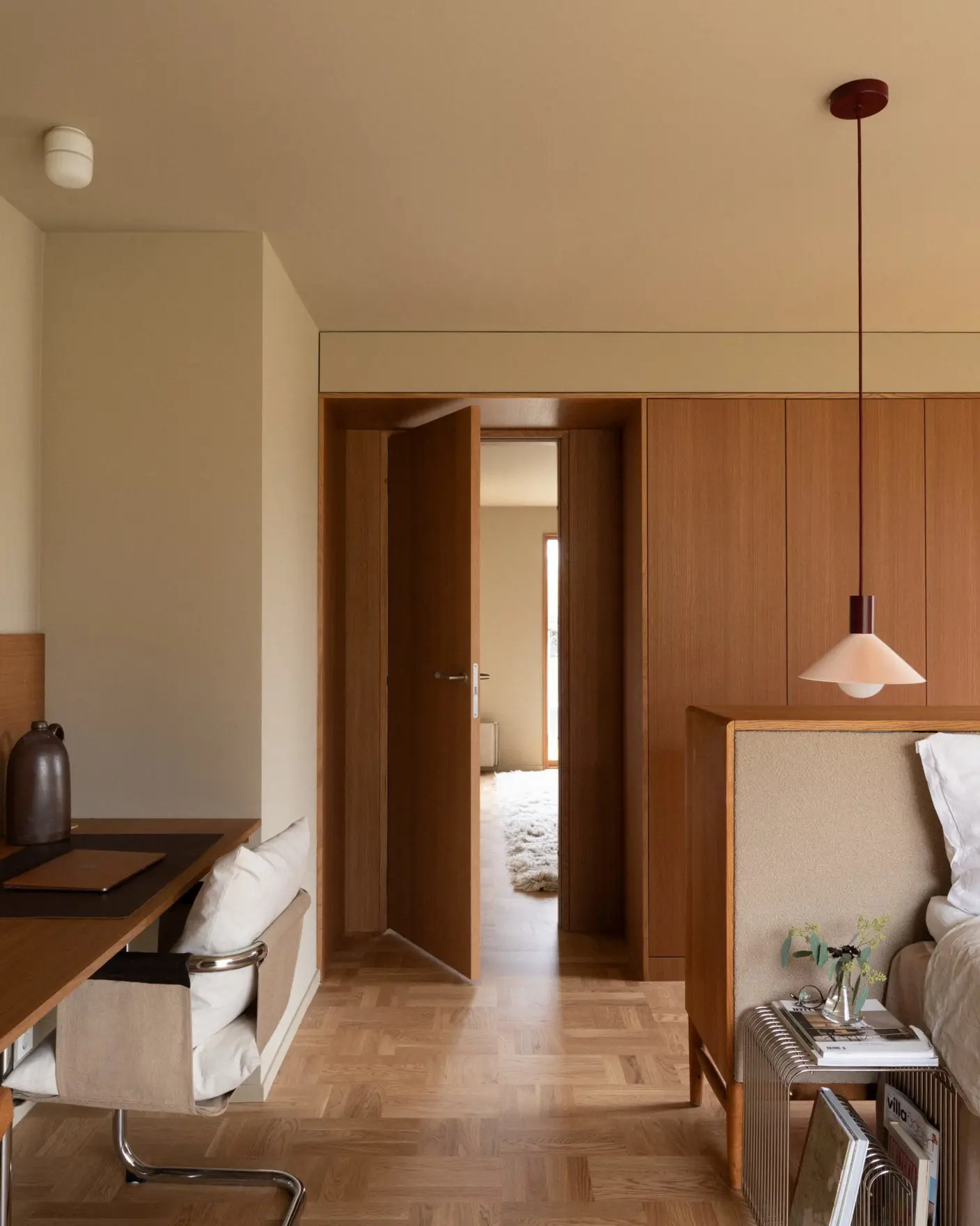 Moderne soverom med tremøbler, åpen dør til annen rom, pendellampe, skrivebord med stol, og sengebenk med dekorative bøker