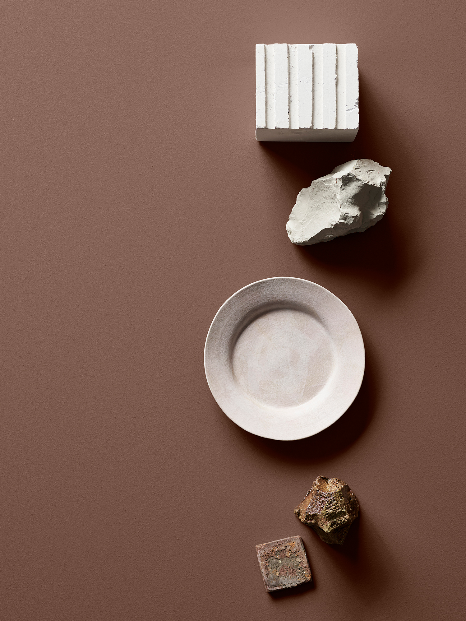 Keramikföremål och stenar arrangerade mot en brun bakgrund, inklusive en vit fyrkantig och en rund platta. Bordsytan är målad i Jotun 20162 Mellow.