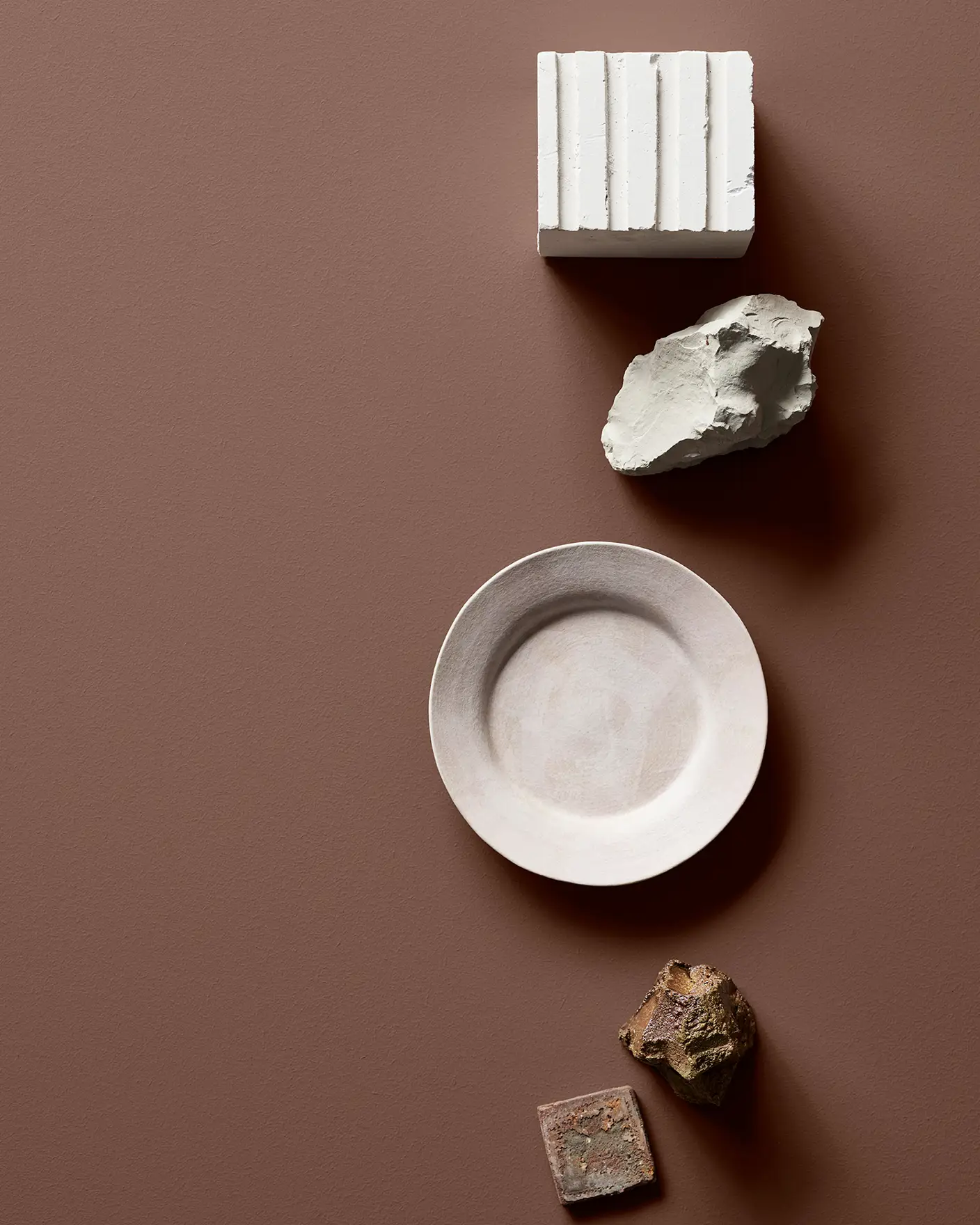 Keramikföremål och stenar arrangerade mot en brun bakgrund, inklusive en vit fyrkantig och en rund platta. Bordsytan är målad i Jotun 20162 Mellow.