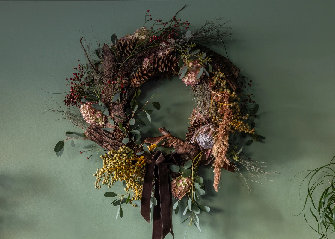 Stor, rund krans med tørkede strå og detaljer i rosa, brunt fløyelsbånd, hengende på grønn vegg i Jotun 7040 Cypress
