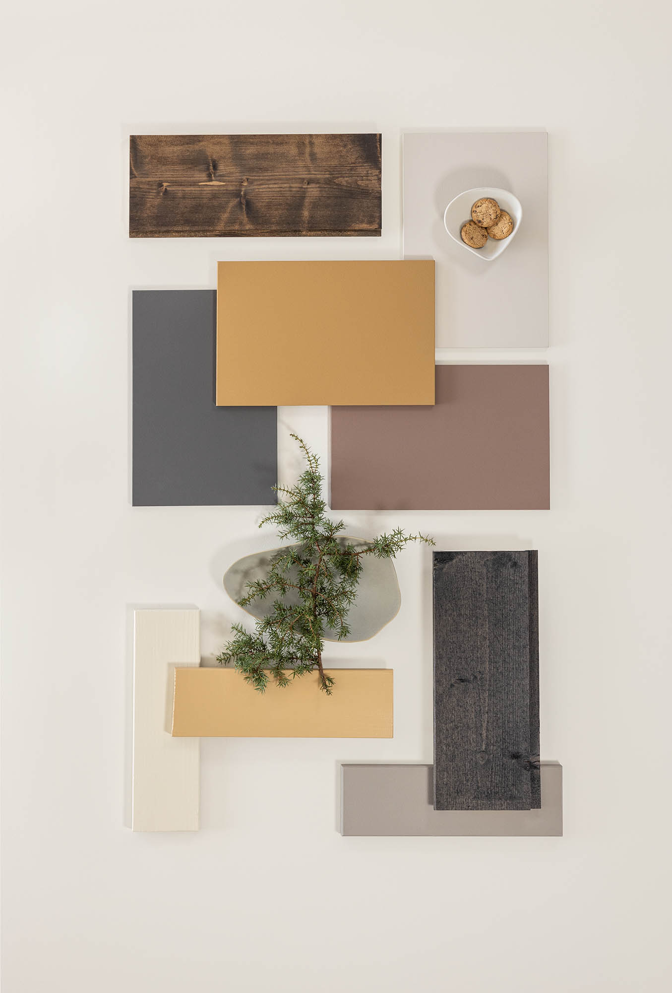 Fargeprøver arrangert på et bord, inkludert ulike nyanser av brunt, grått og hvitt, med en liten hvit skål med kjeks og en grønn kvist for dekorasjon