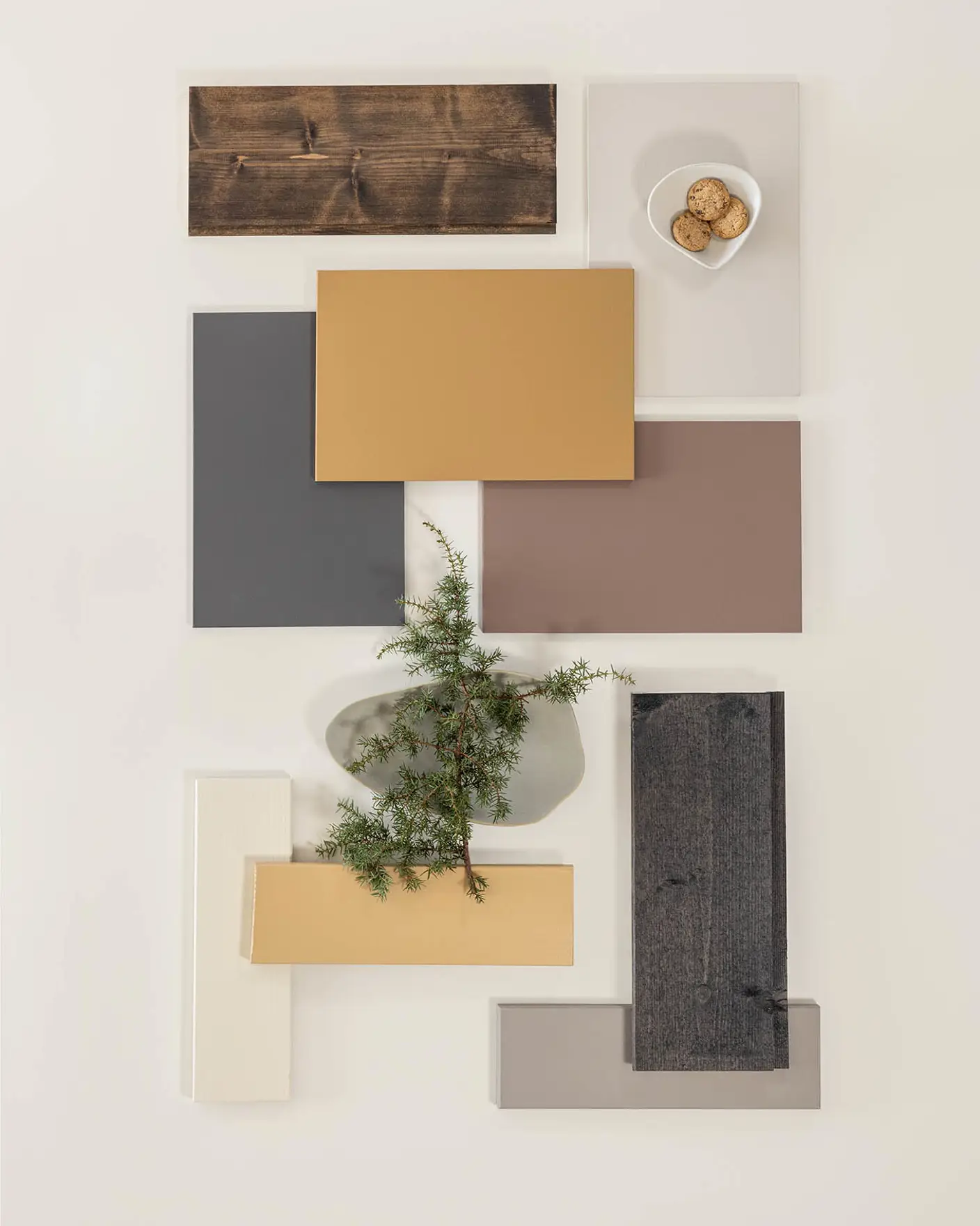 Fargeprøver arrangert på et bord, inkludert ulike nyanser av brunt, grått og hvitt, med en liten hvit skål med kjeks og en grønn kvist for dekorasjon